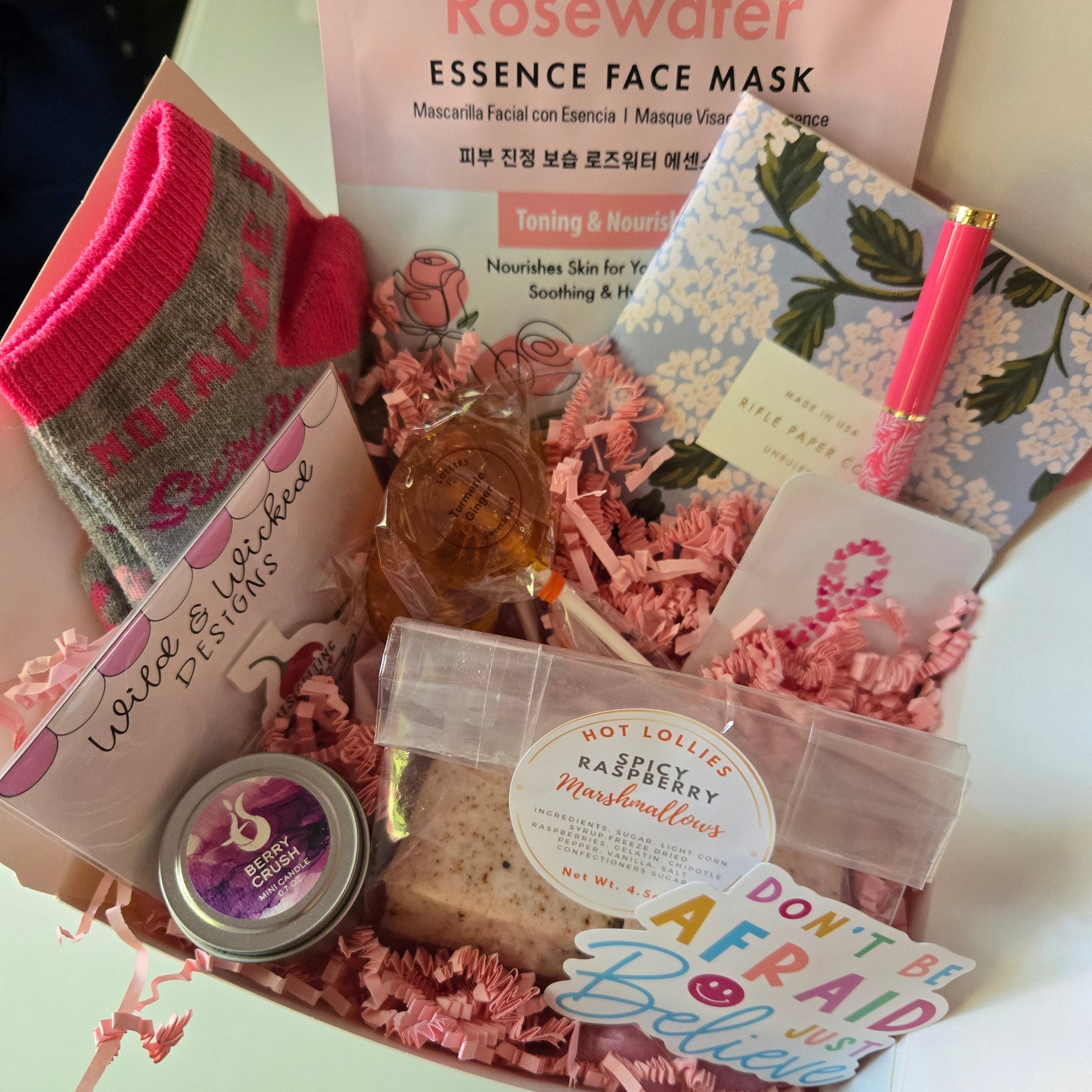 Assorted beauty products including a face mask, candle, and note pad in a box with decorative pink elements.